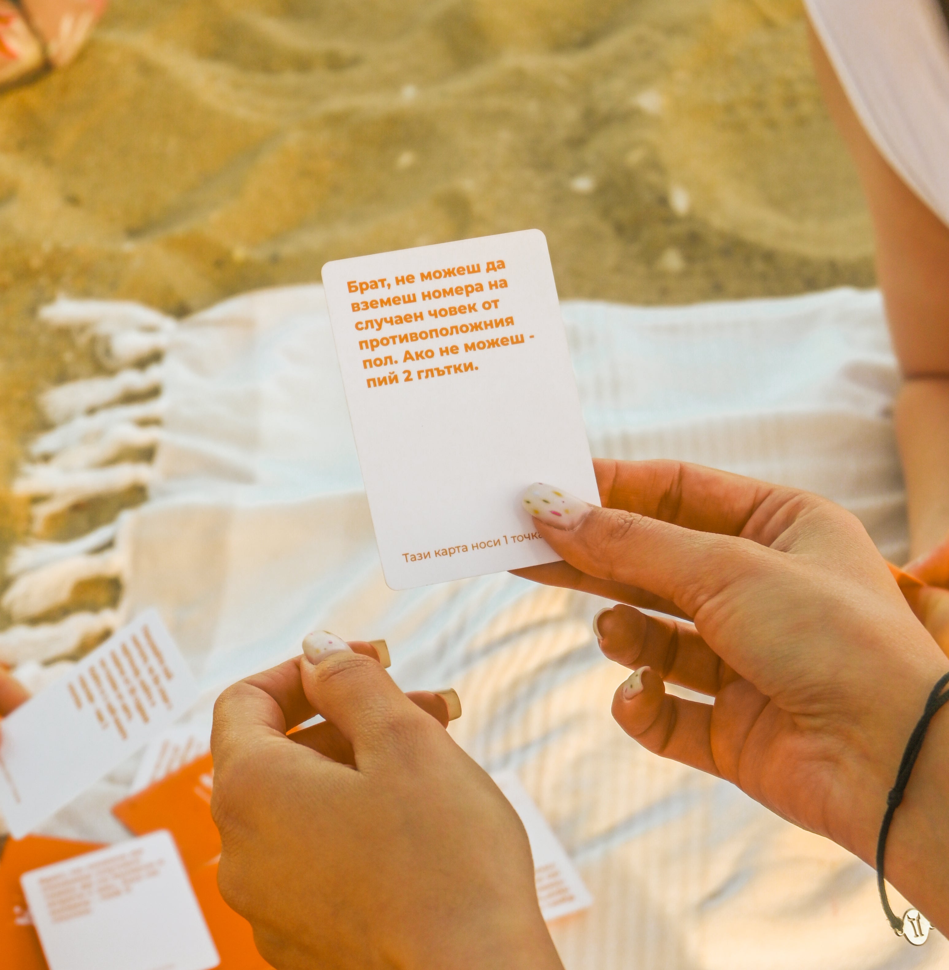 Хора играят парти игра "Брат, не можеш – beach edition" на плаж, държат карта с предизвикателство, а около тях са разпръснати други карти върху хавлия на пясъка.
