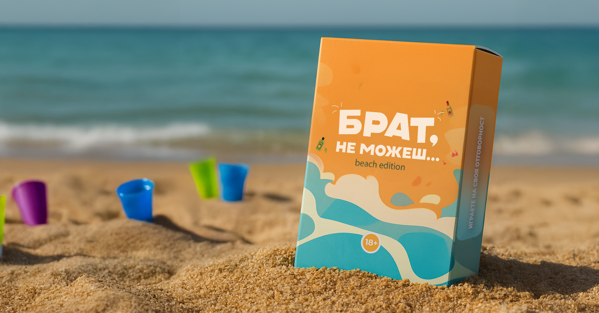 Кутия на парти играта 'Брат, не можеш... Beach Edition', поставена върху пясъка на плаж с море на заден план и цветни кофички около нея.